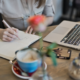 A girl writing on a notebook with a laptop and a cup of coffee