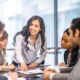 A group of adult men and women are indoors in an office having a meeting.