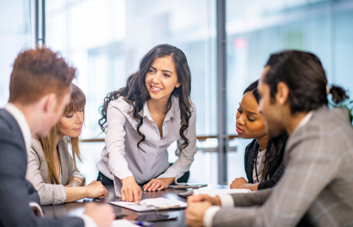 A group of adult men and women are indoors in an office having a meeting.